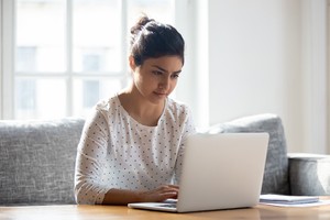 Focused Indian woman using laptop at home, looking at screen, chatting, reading or writing email, sitting on couch, serious female student doing homework, working on research project online